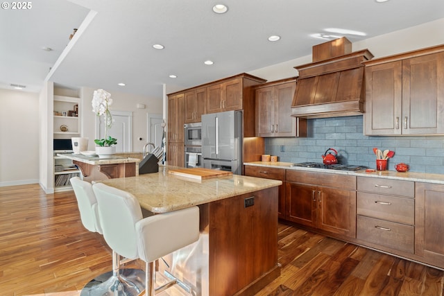 kitchen with dark wood-style flooring, stainless steel appliances, custom range hood, decorative backsplash, and a kitchen breakfast bar