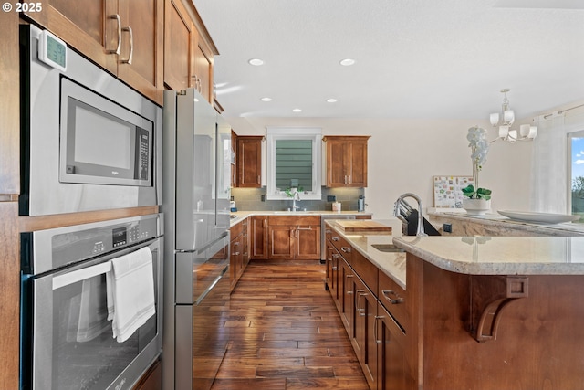 kitchen with tasteful backsplash, appliances with stainless steel finishes, brown cabinetry, dark wood-type flooring, and an island with sink