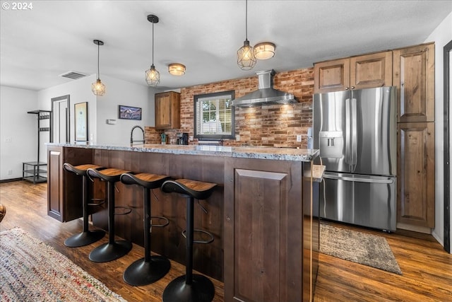 kitchen with light stone counters, visible vents, hanging light fixtures, wall chimney range hood, and stainless steel fridge with ice dispenser