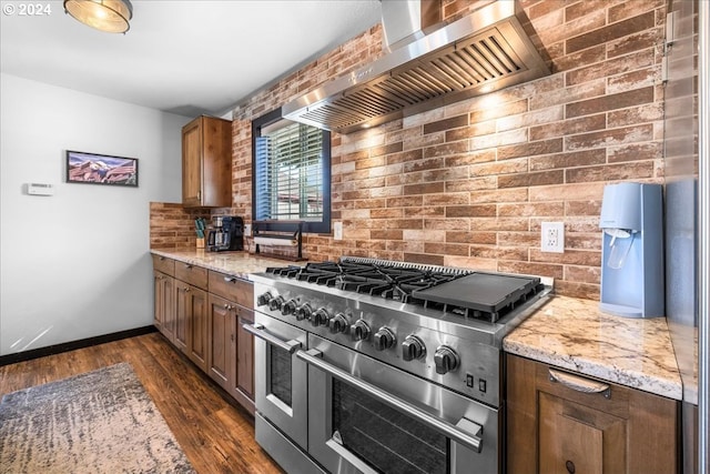 kitchen with light stone counters, dark wood-style floors, backsplash, wall chimney range hood, and double oven range