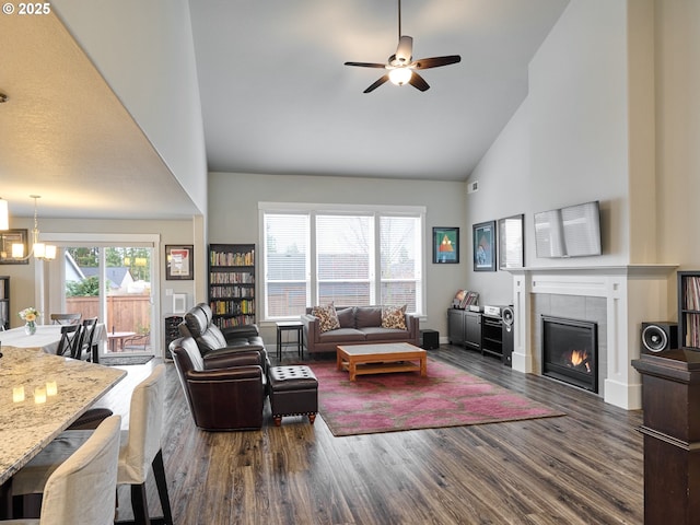 living area featuring high vaulted ceiling, a tiled fireplace, ceiling fan with notable chandelier, baseboards, and dark wood-style flooring