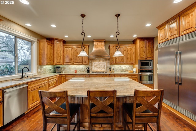 kitchen with a kitchen island, sink, hanging light fixtures, built in appliances, and custom range hood