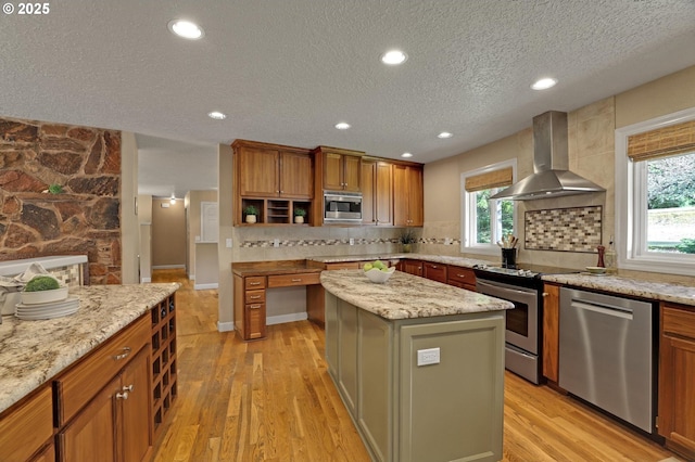 kitchen with a kitchen island, light wood-style floors, appliances with stainless steel finishes, wall chimney exhaust hood, and tasteful backsplash