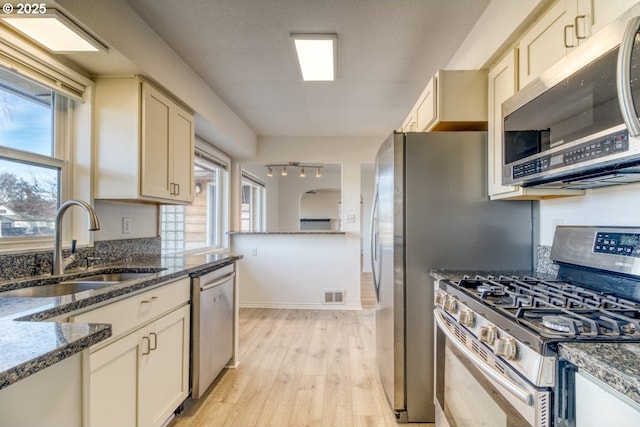 kitchen with sink, appliances with stainless steel finishes, light hardwood / wood-style floors, cream cabinetry, and dark stone counters