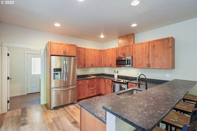 kitchen featuring stainless steel appliances, a sink, a peninsula, light wood finished floors, and dark stone counters