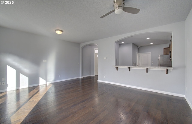 unfurnished living room featuring a textured ceiling, arched walkways, baseboards, ceiling fan, and dark wood-style flooring