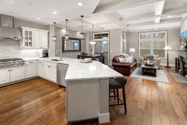 kitchen with appliances with stainless steel finishes, a sink, wall chimney range hood, coffered ceiling, and a peninsula