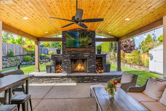 view of patio / terrace with ceiling fan, an outdoor living space with a fireplace, an outdoor structure, a fenced backyard, and a storage unit