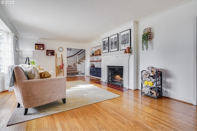 living room featuring light wood-style flooring, a fireplace with flush hearth, baseboards, stairs, and crown molding