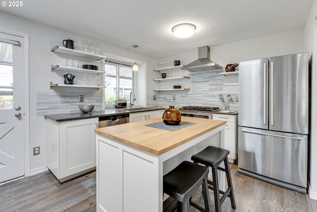 kitchen with open shelves, wooden counters, wall chimney range hood, a sink, and stainless steel appliances