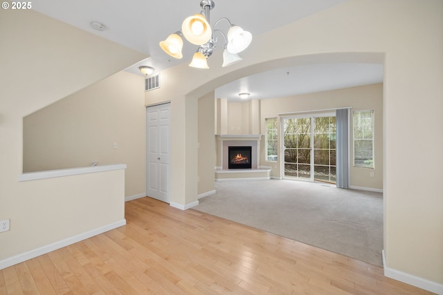 unfurnished living room with visible vents, baseboards, a warm lit fireplace, an inviting chandelier, and wood finished floors