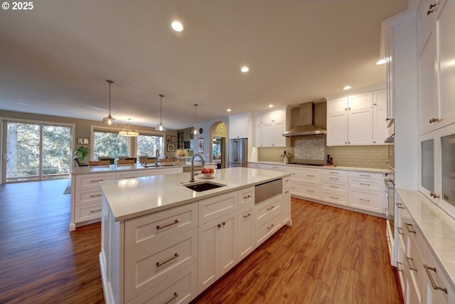 kitchen with an island with sink, wall chimney range hood, hanging light fixtures, stainless steel refrigerator, and sink