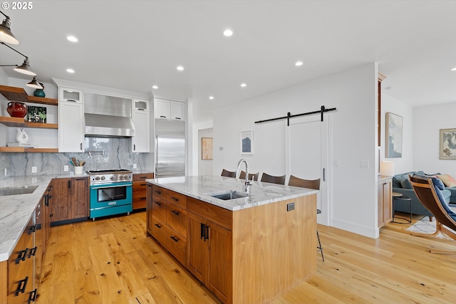 kitchen with a barn door, premium appliances, light wood finished floors, a sink, and wall chimney range hood