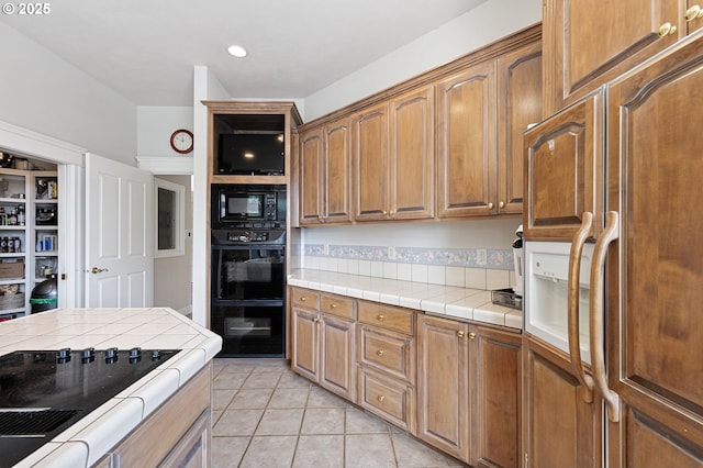 kitchen featuring brown cabinetry, black appliances, tile counters, and light tile patterned flooring