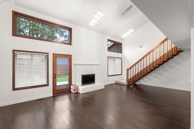 unfurnished living room with a brick fireplace, dark wood finished floors, stairway, an inviting chandelier, and high vaulted ceiling