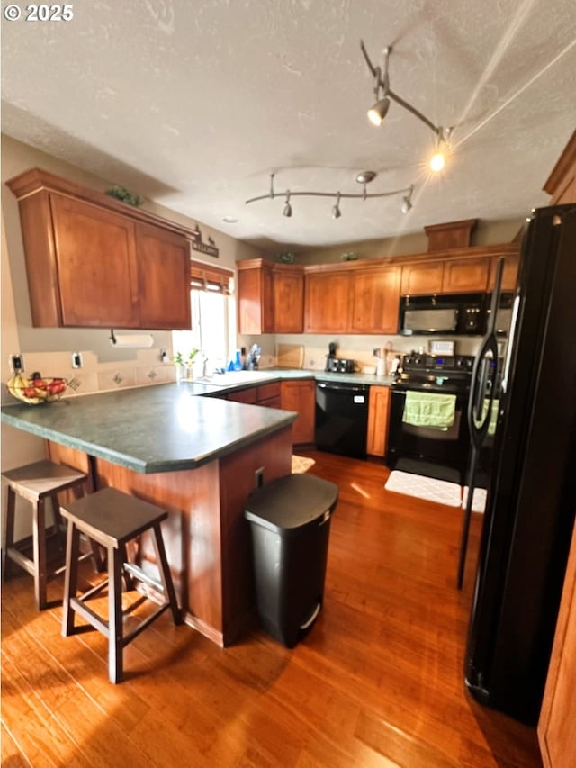 kitchen featuring brown cabinets, black appliances, a textured ceiling, wood finished floors, and a peninsula