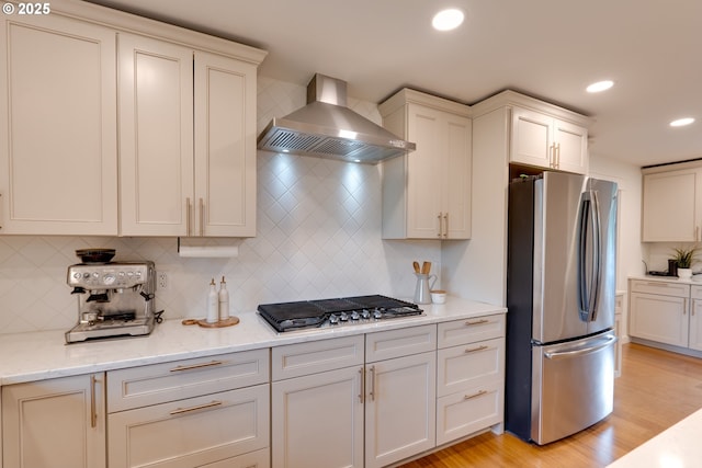 kitchen with stainless steel appliances, light stone counters, wall chimney range hood, light hardwood / wood-style floors, and backsplash