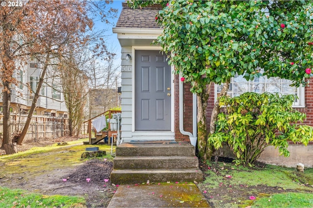 entrance to property featuring brick siding, a shingled roof, and fence