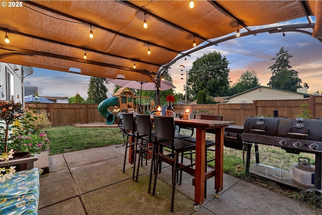 patio terrace at dusk featuring a lawn, a fenced backyard, and a playground