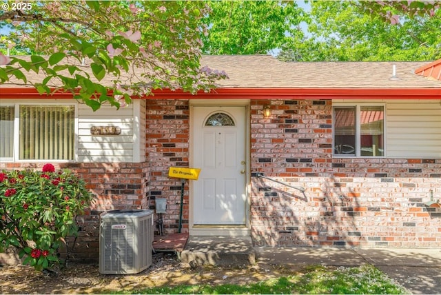 doorway to property featuring central air condition unit, brick siding, and a shingled roof