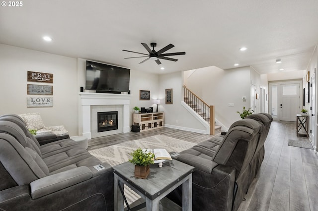 living area featuring a glass covered fireplace, wood finished floors, stairway, recessed lighting, and a ceiling fan