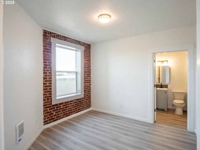 unfurnished bedroom featuring ensuite bathroom, brick wall, light wood-type flooring, and baseboards