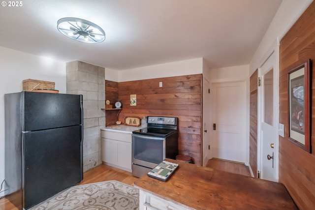 kitchen featuring black refrigerator, stainless steel range with electric cooktop, light hardwood / wood-style floors, white cabinets, and wooden walls