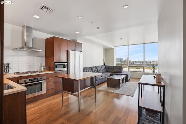 kitchen featuring wall chimney range hood, appliances with stainless steel finishes, decorative backsplash, light wood-type flooring, and visible vents
