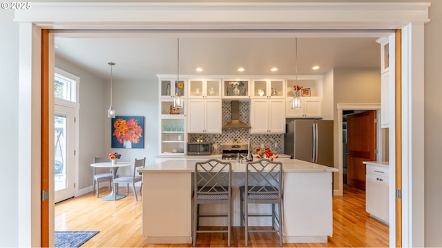 kitchen featuring pendant lighting, appliances with stainless steel finishes, white cabinets, a center island with sink, and wall chimney exhaust hood