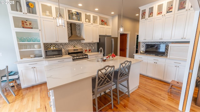 kitchen with appliances with stainless steel finishes, sink, wall chimney range hood, hanging light fixtures, and a center island with sink