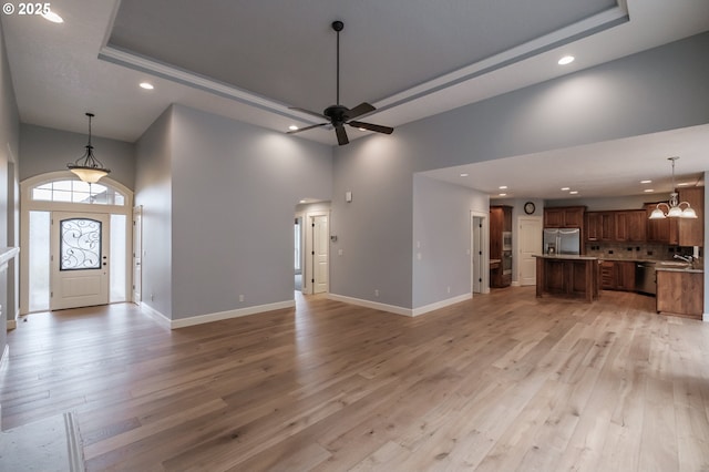 unfurnished living room with a towering ceiling, ceiling fan with notable chandelier, sink, a tray ceiling, and light wood-type flooring