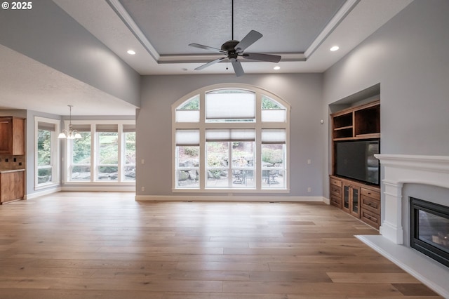 unfurnished living room featuring a tray ceiling, ceiling fan with notable chandelier, light hardwood / wood-style floors, and a textured ceiling