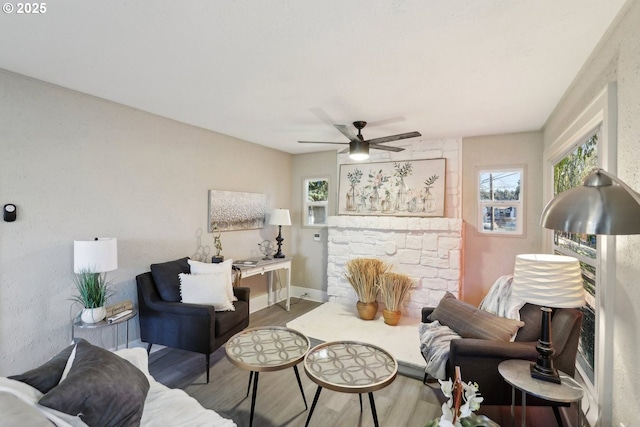 living area with ceiling fan, a stone fireplace, baseboards, and wood finished floors