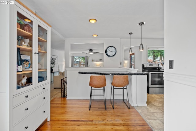 kitchen with a kitchen breakfast bar, a ceiling fan, dark countertops, stainless steel electric stove, and white cabinetry