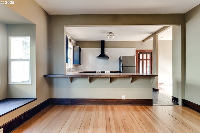 kitchen featuring wall chimney exhaust hood, light hardwood / wood-style flooring, stainless steel fridge, kitchen peninsula, and a kitchen breakfast bar