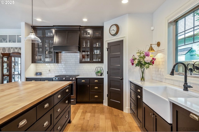 kitchen with custom exhaust hood, wooden counters, stainless steel gas range oven, tasteful backsplash, and a sink