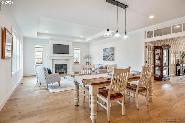 dining space with a glass covered fireplace, baseboards, light wood-type flooring, recessed lighting, and a raised ceiling