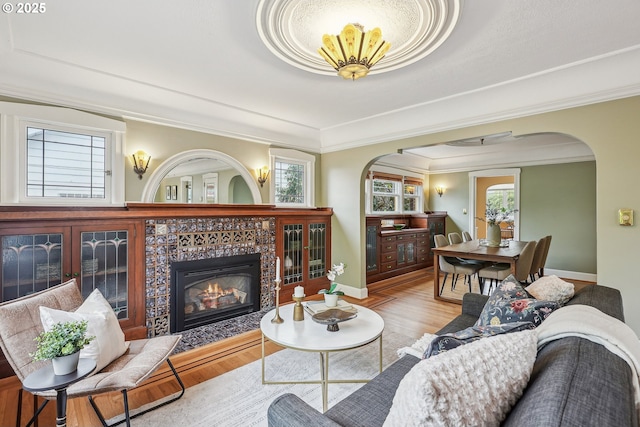 living area featuring crown molding, wood finished floors, baseboards, a tiled fireplace, and arched walkways