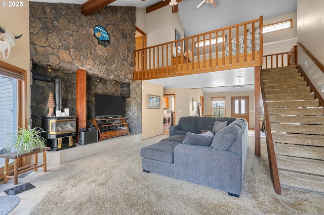 carpeted living area featuring beamed ceiling, a high ceiling, stairs, ceiling fan, and a wood stove