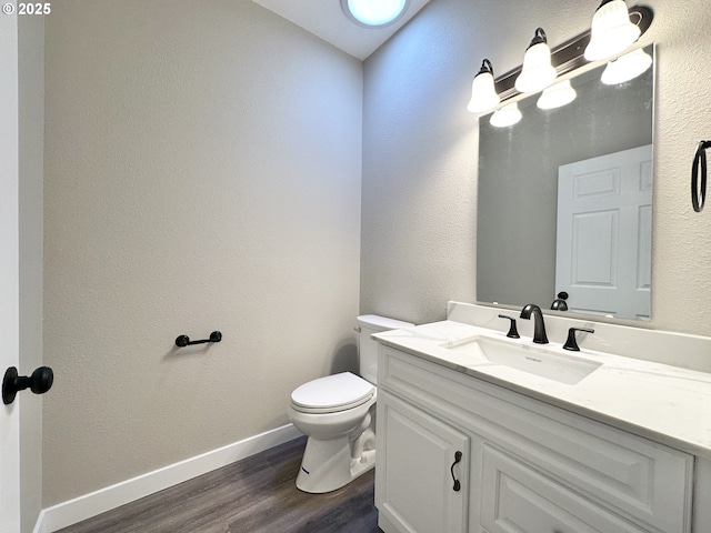 half bathroom with toilet, wood finished floors, baseboards, vanity, and a textured wall