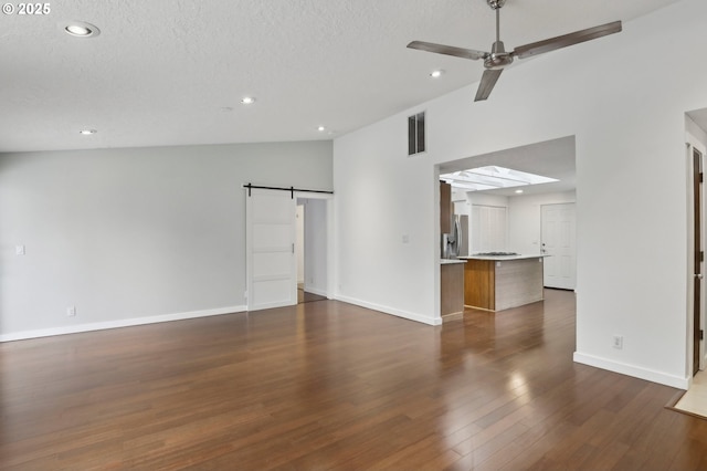 unfurnished living room featuring a ceiling fan, visible vents, dark wood finished floors, vaulted ceiling with skylight, and a barn door