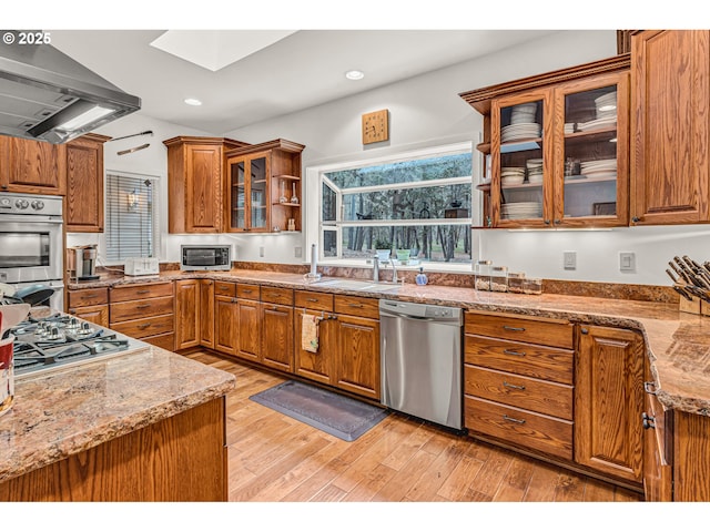 kitchen with wall chimney range hood, stainless steel appliances, brown cabinetry, and a sink