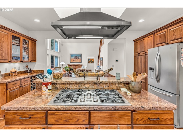 kitchen featuring brown cabinets, light stone counters, wall chimney exhaust hood, stainless steel appliances, and glass insert cabinets