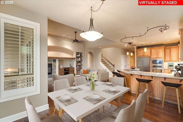 dining room with a ceiling fan, baseboards, dark wood finished floors, stairs, and a tiled fireplace