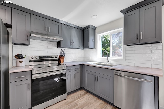 kitchen featuring appliances with stainless steel finishes, under cabinet range hood, a sink, gray cabinetry, and light countertops