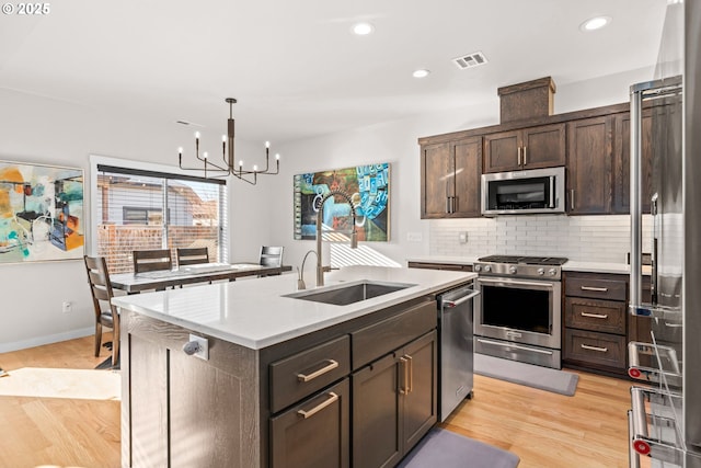 kitchen featuring dark brown cabinetry, a center island with sink, stainless steel appliances, light hardwood / wood-style floors, and decorative backsplash