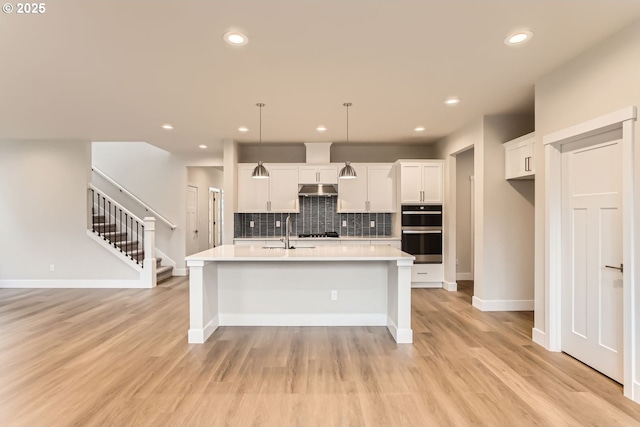 kitchen featuring a center island with sink, stainless steel double oven, white cabinetry, and hanging light fixtures