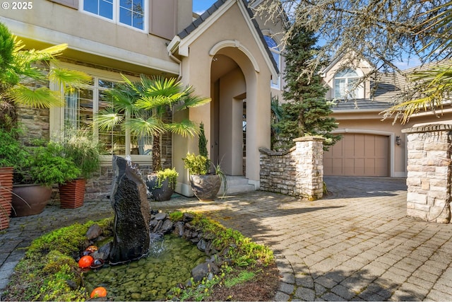 view of exterior entry with decorative driveway, stone siding, a garage, and stucco siding