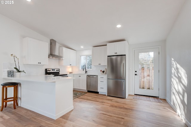 kitchen with a peninsula, a sink, appliances with stainless steel finishes, light wood-type flooring, and wall chimney exhaust hood