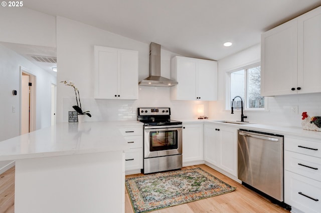 kitchen featuring light wood finished floors, appliances with stainless steel finishes, a sink, a peninsula, and wall chimney exhaust hood